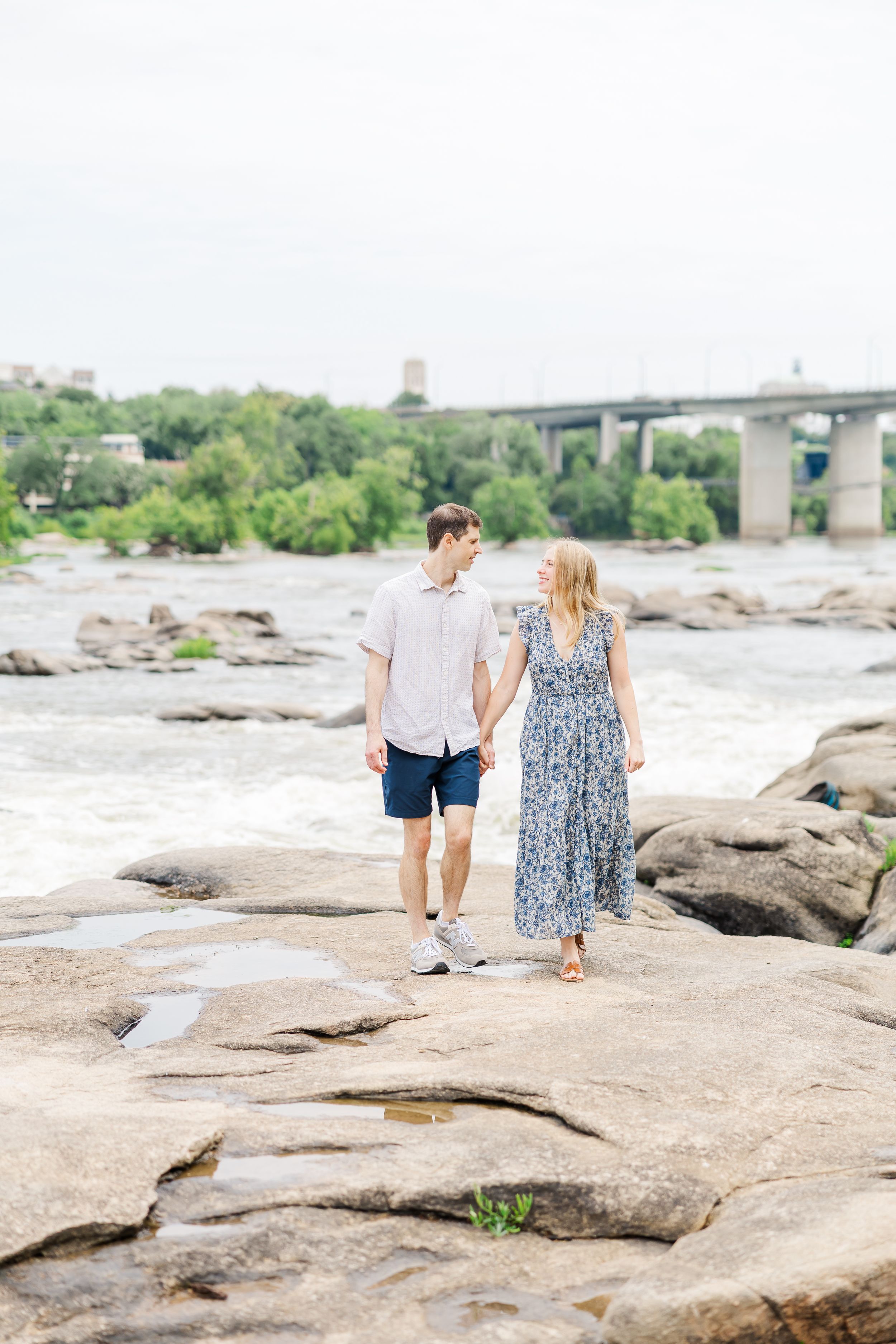 Couple holding hands walking on rocky riverbank with bridge and lush greenery in background on overcast day.