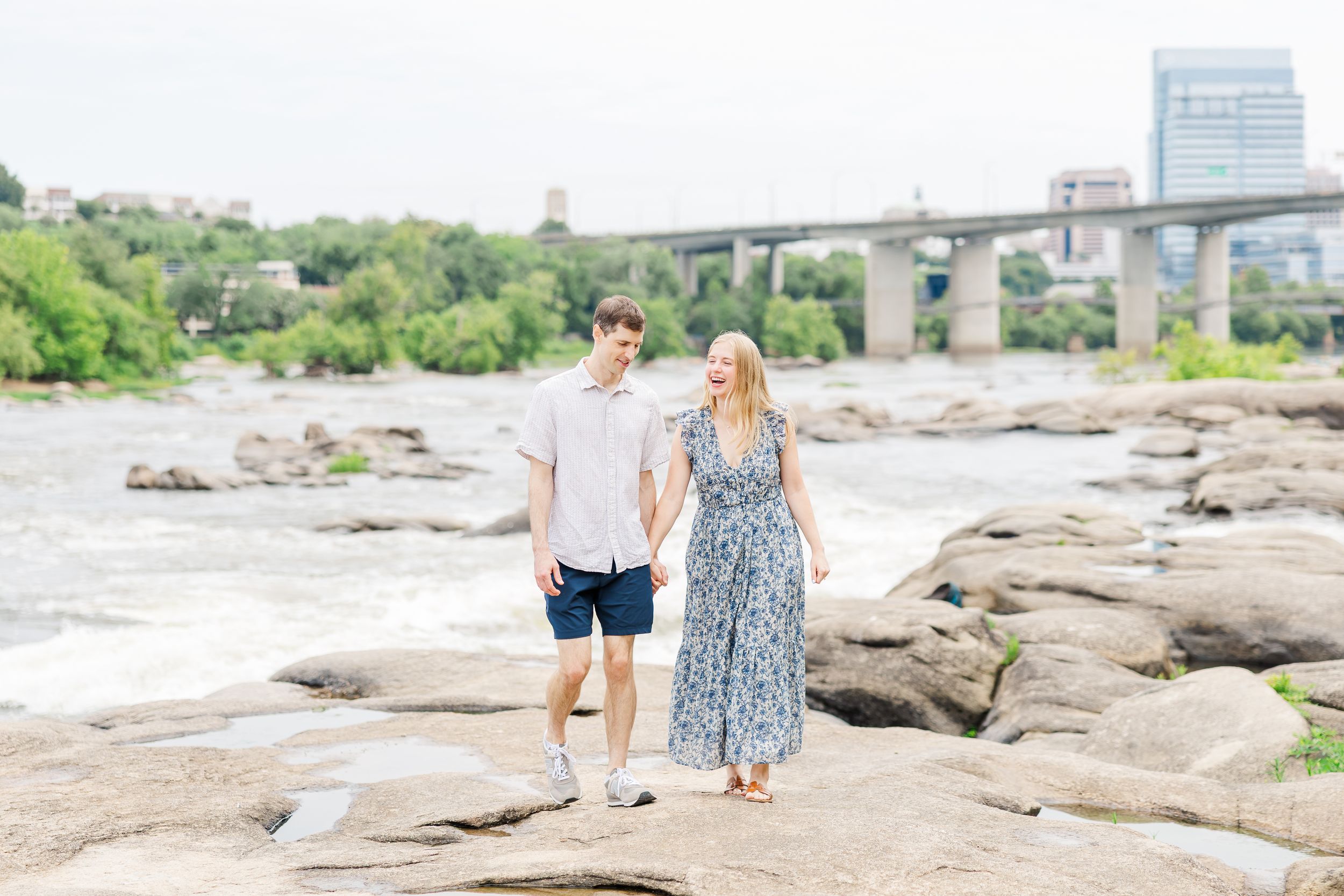 Couple holding hands walking on rocks by a river with a bridge and city skyline in the background.