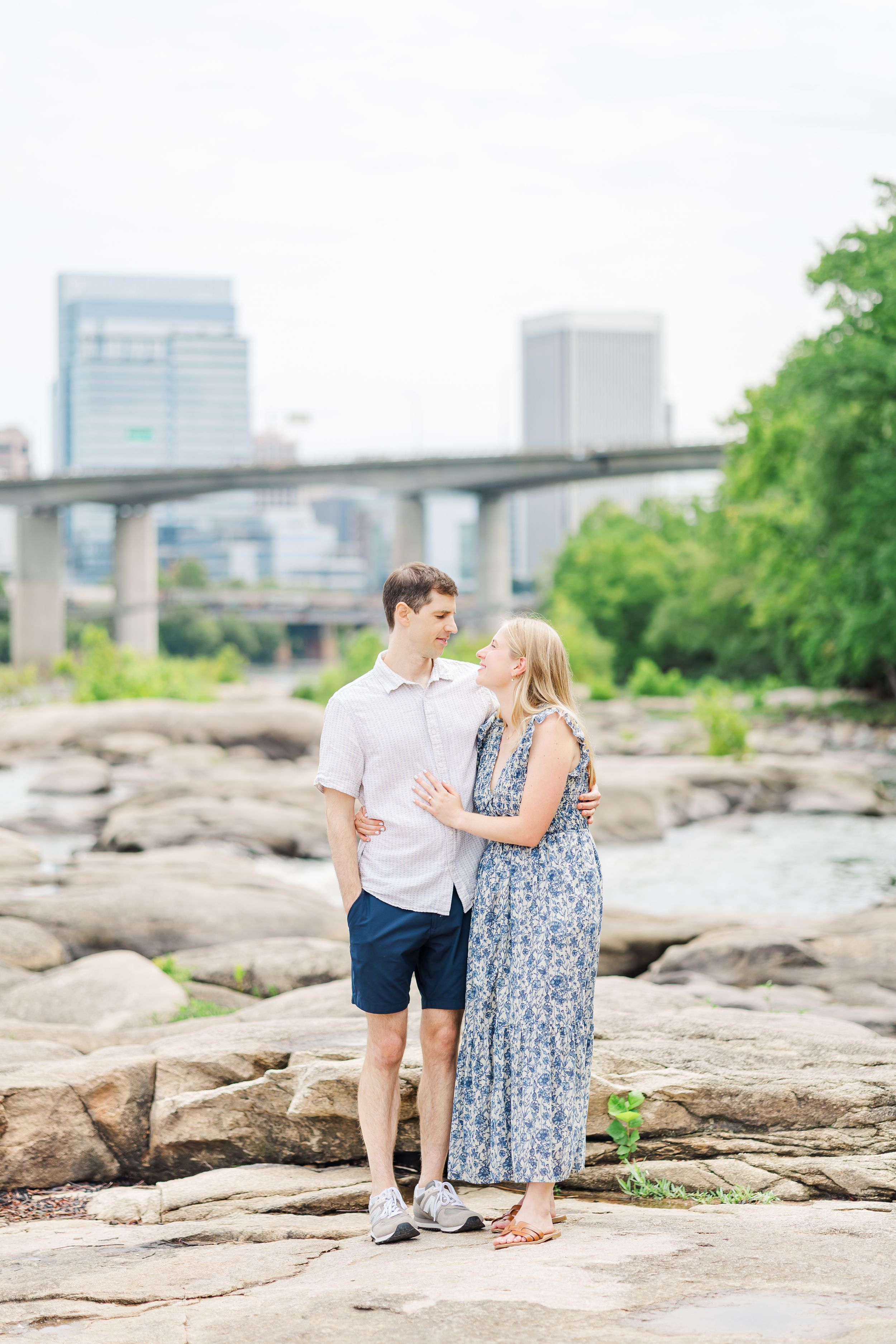 Couple embracing on rocky riverbank with city skyline and bridge in background on a sunny day.