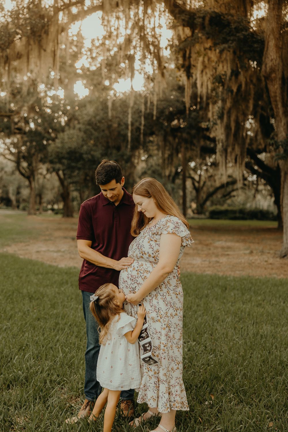 A pregnant woman in a floral dress stands with her husband and toddler daughter in a sunlit park with moss-draped oak trees.