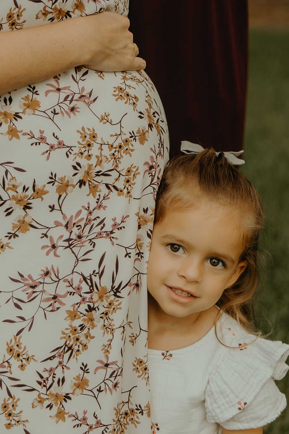 A young girl in a white dress peeks out from behind a pregnant woman in a floral dress outdoors.