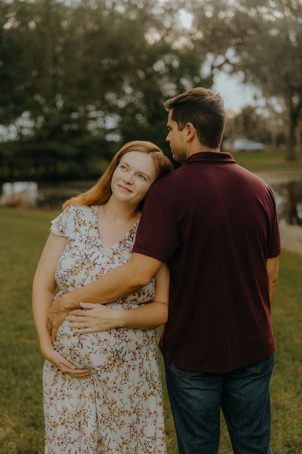 Pregnant woman in floral dress embraced by partner outdoors during golden hour maternity photo session.
