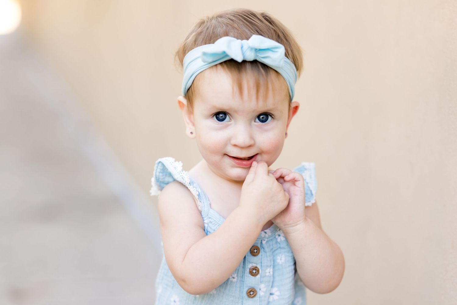 Adorable toddler girl with blue eyes wearing a light blue romper and bow headband, smiling with hands clasped.