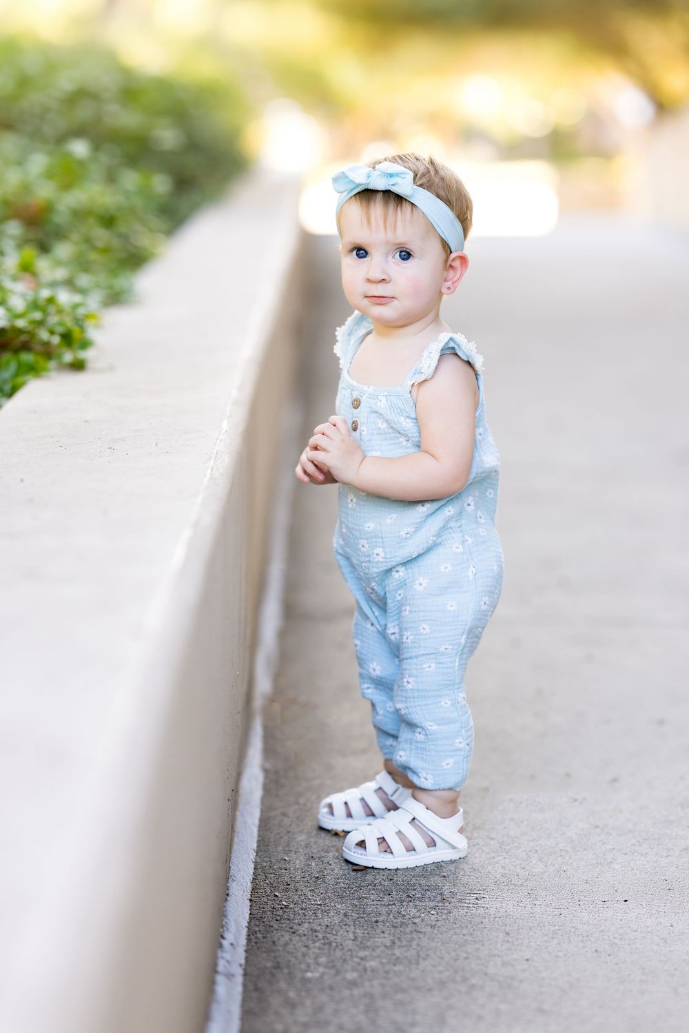 Adorable baby girl in light blue romper and bow headband standing on a sunny sidewalk wearing white sandals.