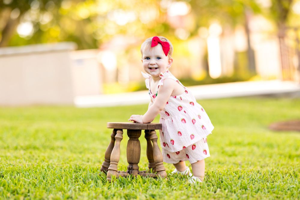 Smiling baby girl in white dress with red bow stands at wooden stool on green grass outdoors.