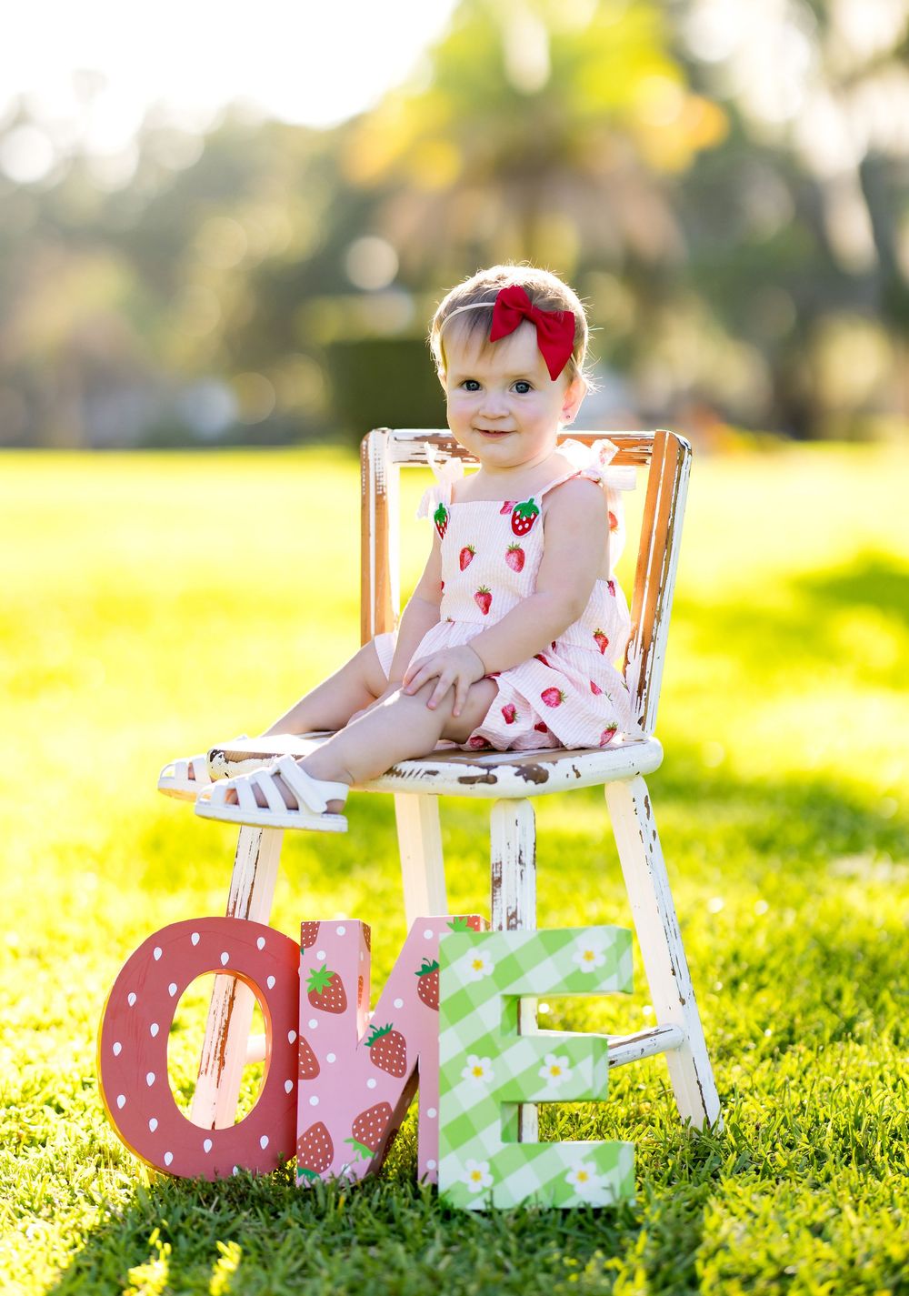 Baby girl in strawberry romper sits on white chair outdoors beside colorful ONE letters for first birthday photos.