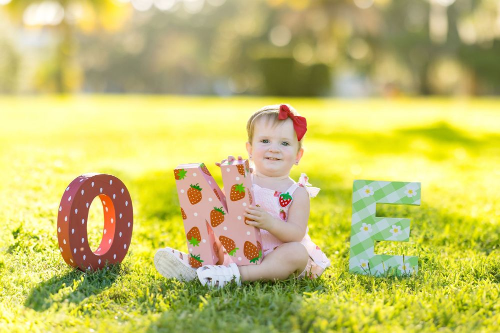 Baby girl in pink strawberry dress sits on grass with decorative ONE letters for her first birthday photo.