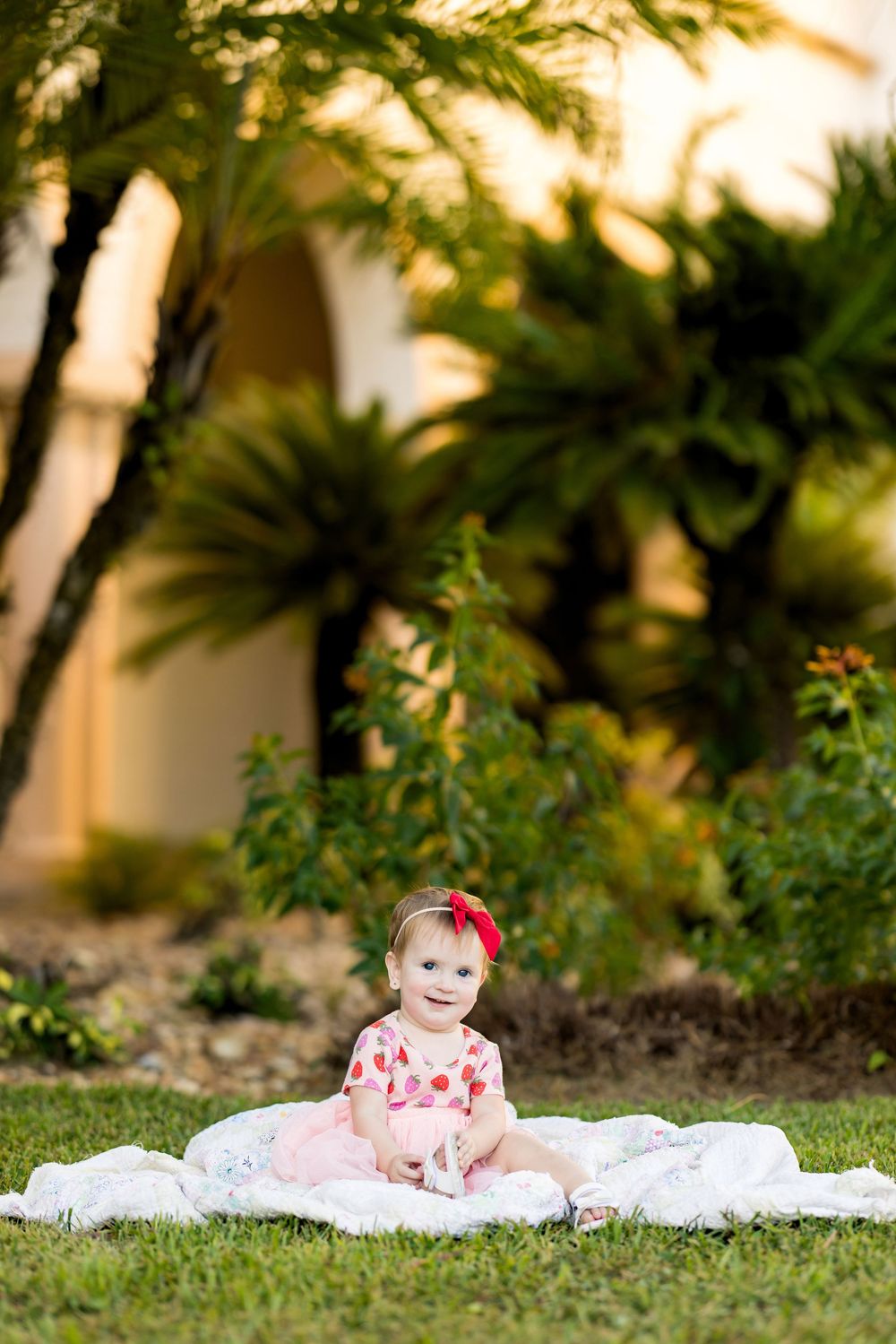 A cute baby girl in a pink floral outfit sits on a blanket in a lush green garden with palm trees.