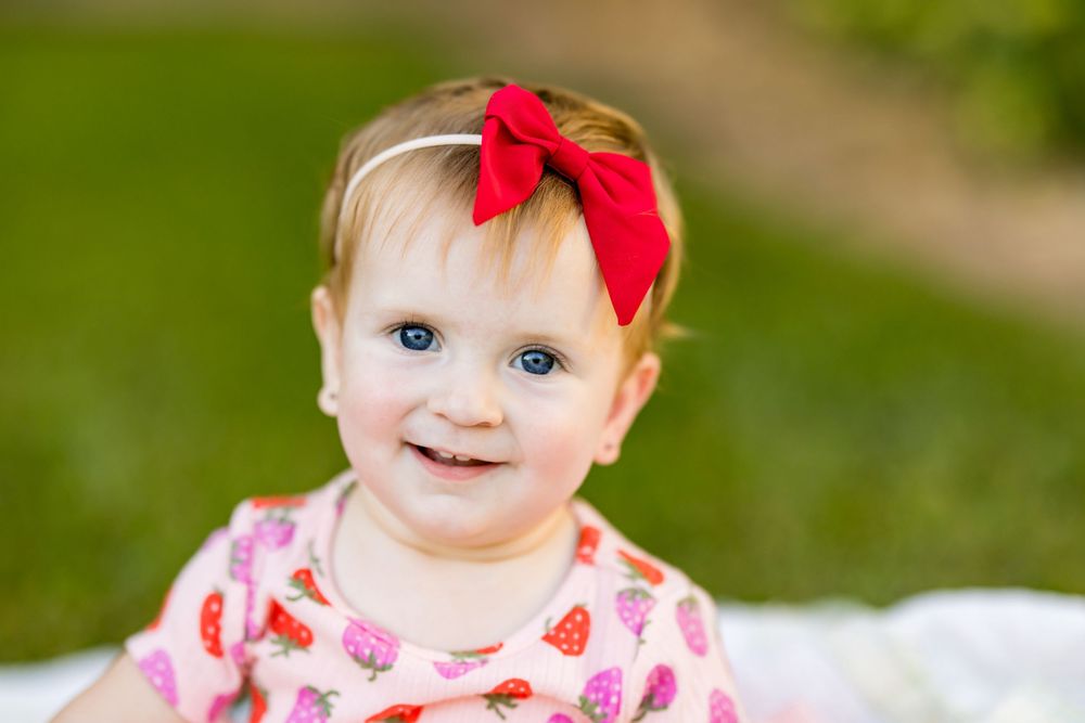 Smiling baby girl with blue eyes wearing a red bow headband and pink strawberry dress outdoors.