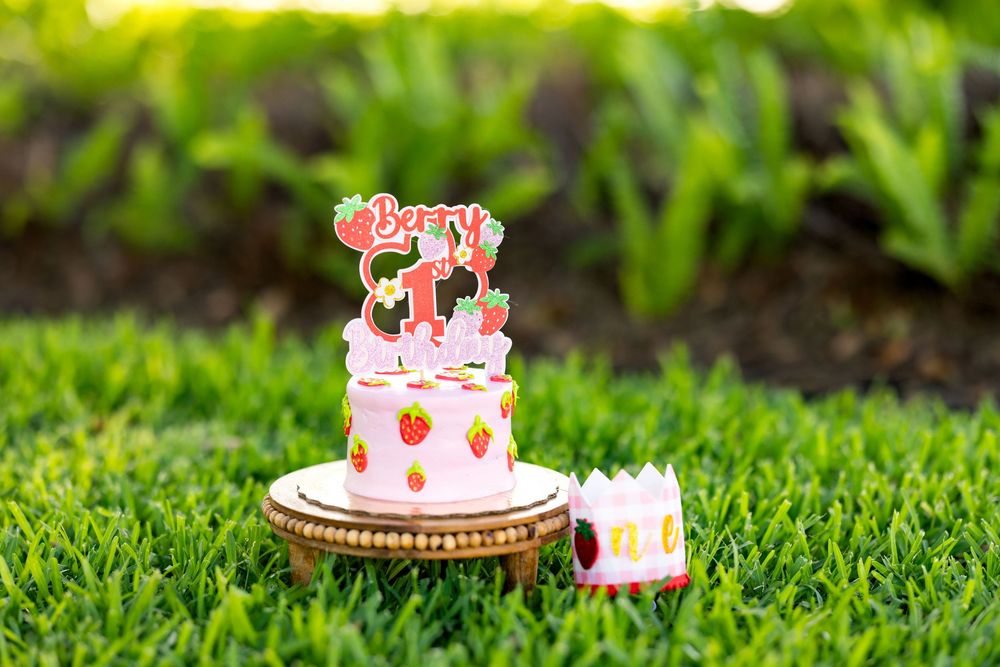 A two-tier strawberry first birthday cake with Berry 1 topper on a wooden stand in green grass.