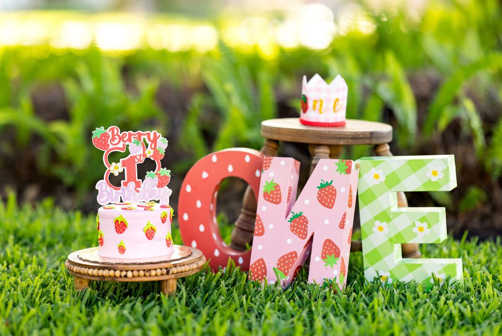 Strawberry-themed first birthday party decorations spelling ONE with pink and green letters, a smash cake, and crown on green grass.
