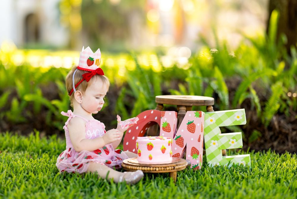 Baby girl in strawberry crown celebrates 1st birthday with cake on green grass outdoors.