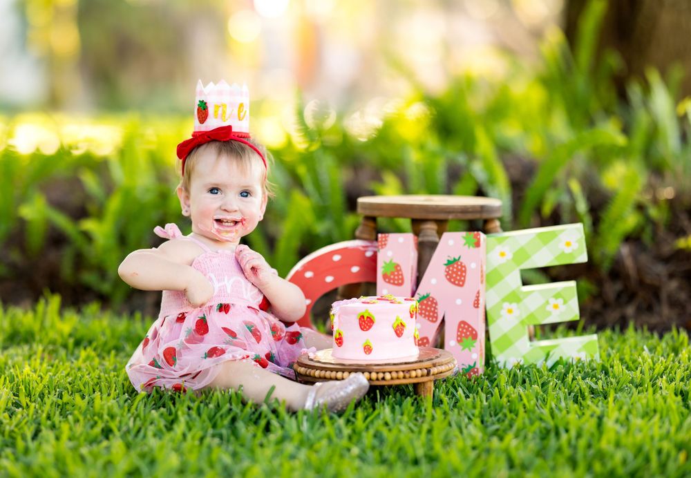 Baby girl in strawberry dress and crown sits on grass celebrating her first birthday with decorative letters and cake.