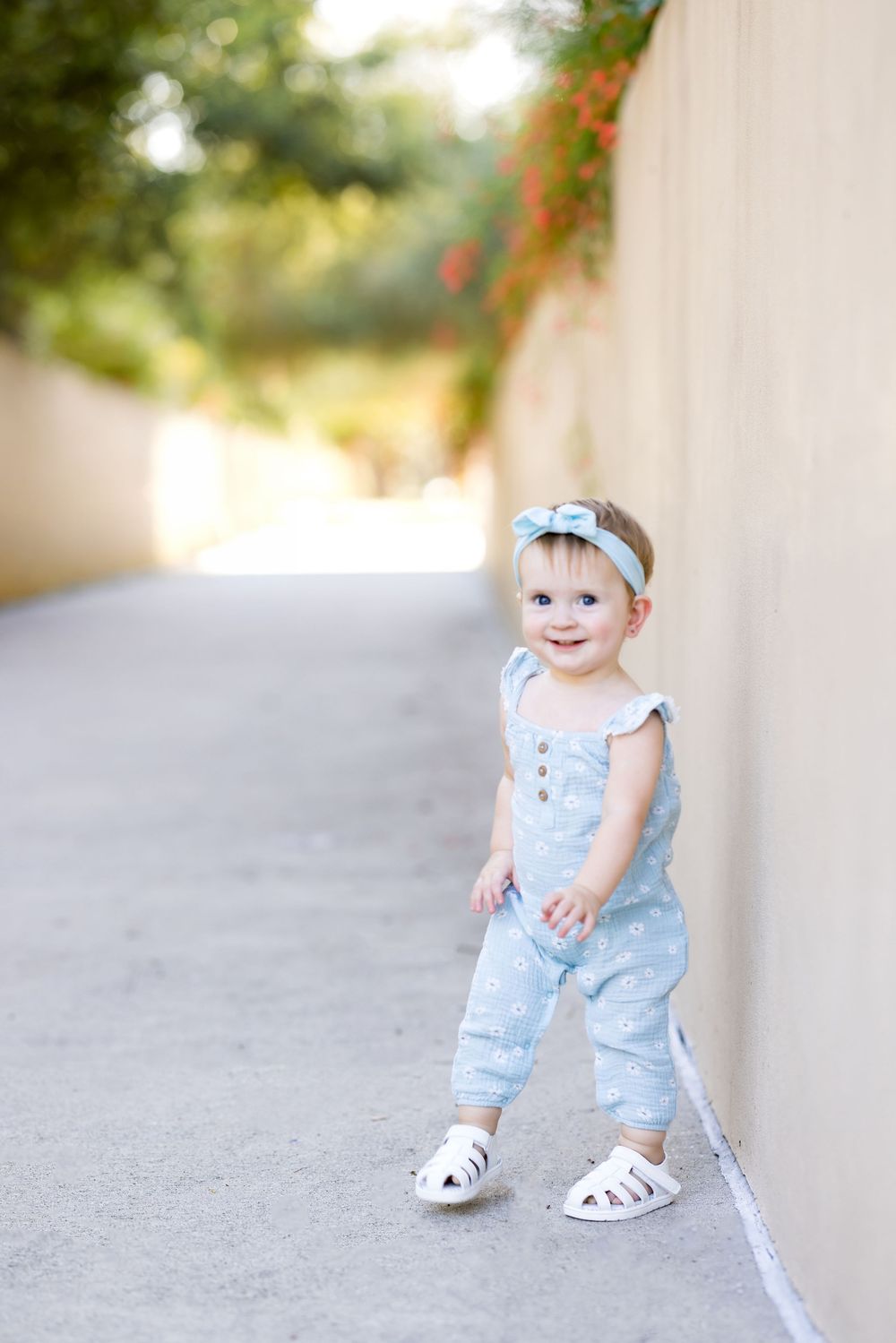 Adorable baby girl in light blue romper and bow headband smiling while standing in a sunny outdoor pathway.