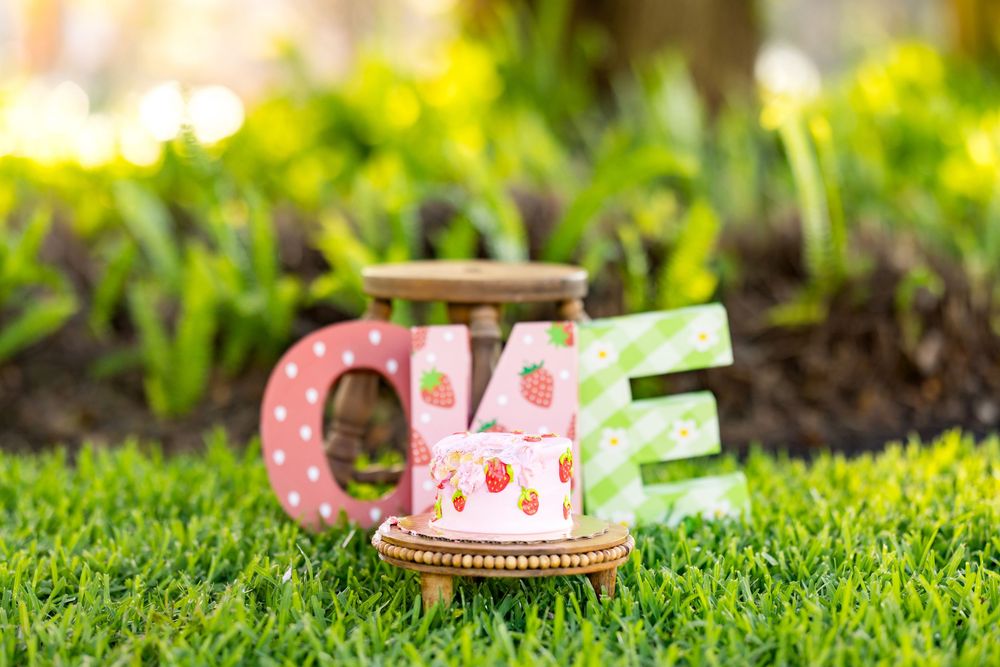 A small strawberry-decorated first birthday cake sits on grass beside colorful ONE letters in a sunny garden setting.