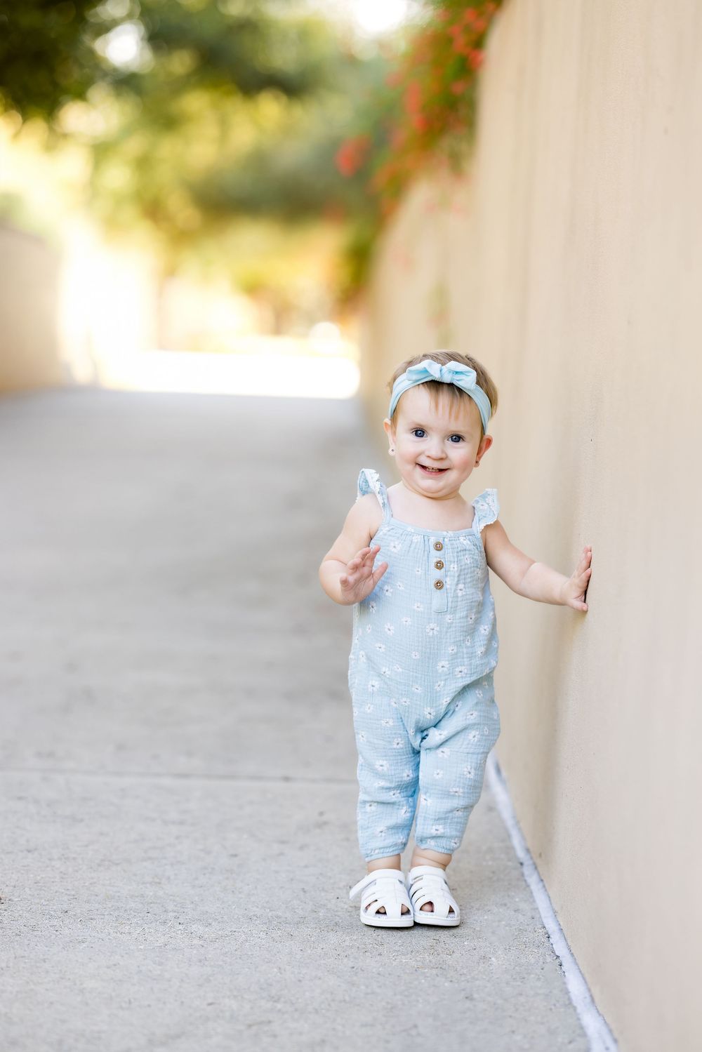 Smiling baby girl in light blue floral romper and bow headband walking along a sunny outdoor pathway.