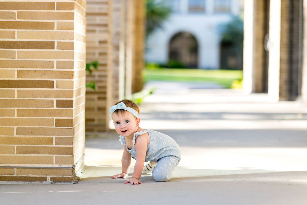 Adorable baby girl in gray romper crawling on outdoor pathway beside brick wall in sunny courtyard.