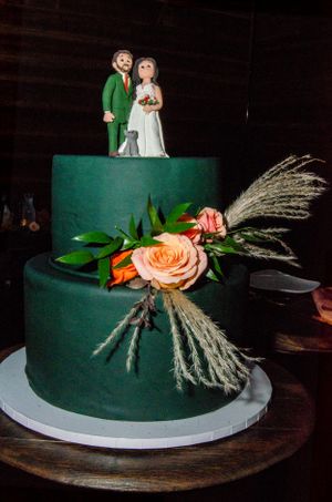 Elegant dark green wedding cake decorated with peach roses and pampas grass, topped with bride and groom figures.