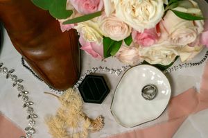 Wedding details flatlay with brown leather shoes, delicate pink roses, and jewelry on white fabric.