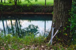A serene pond reflects trees and sky with a sword prop leaning against a tree trunk.