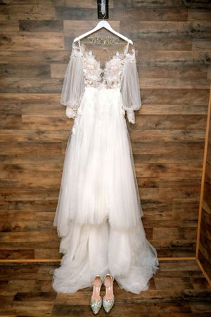 An elegant white wedding dress with lace details and flowing sleeves hangs against a wooden wall with shoes below.