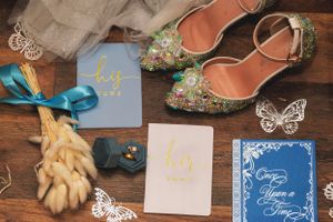 Wedding accessories including sparkly shoes, blue cards, gloves and butterfly decorations arranged on wooden surface.