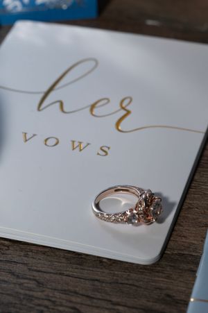 Wedding ring rests on a white card with 'her vows' written in elegant gold lettering.