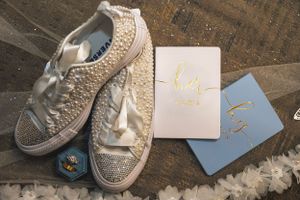 Bejeweled white sneakers and wedding stationery arranged on dark wooden surface.