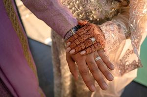 Close-up view of hands wearing traditional Indian bridal jewelry and henna designs against a pink background.