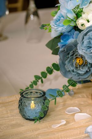 Blue peony floral arrangement with eucalyptus and a blue glass votive candle on a gold table runner.