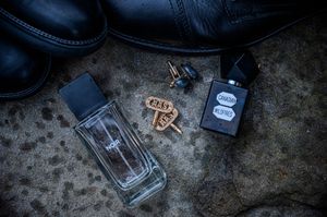 Men's cologne bottles and cufflinks arranged beside black leather shoes on a textured surface.