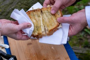 Two people pulling apart a melted grilled cheese sandwich over a wooden cutting board with napkins.