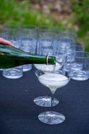 Champagne being poured from a green bottle into a coupe glass on a black table with empty glasses behind.