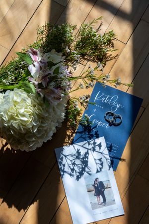 Wedding flat lay with floral bouquet, navy blue invitation suite, rings, and couple photo on wood floor.