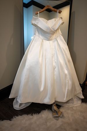Elegant ivory satin off-shoulder ballgown wedding dress on hanger with silver heels on fluffy white rug.