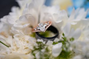 Rose gold diamond engagement ring and black wedding band nestled in white flowers.