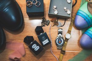 Flat lay of accessories including black dress shoes, cologne, a watch, pearl necklace, earrings, rings, and colorful heels.