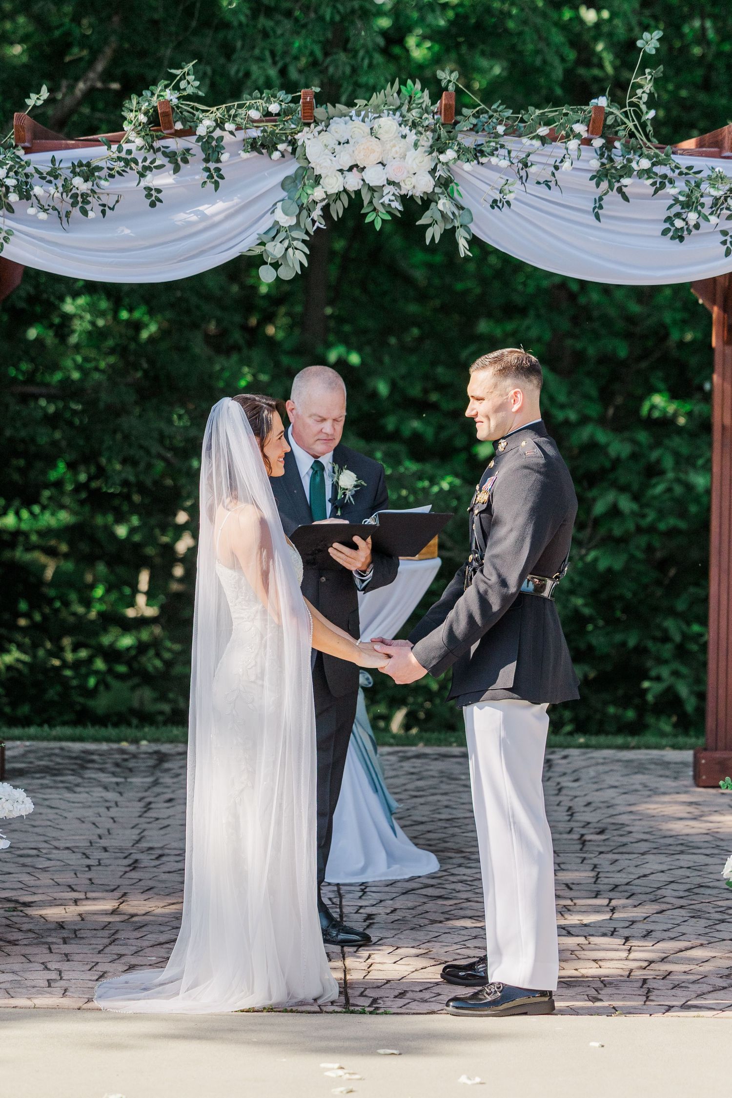Outdoor wedding ceremony with bride in white gown and veil, groom in military dress uniform, exchanging vows under floral arch.