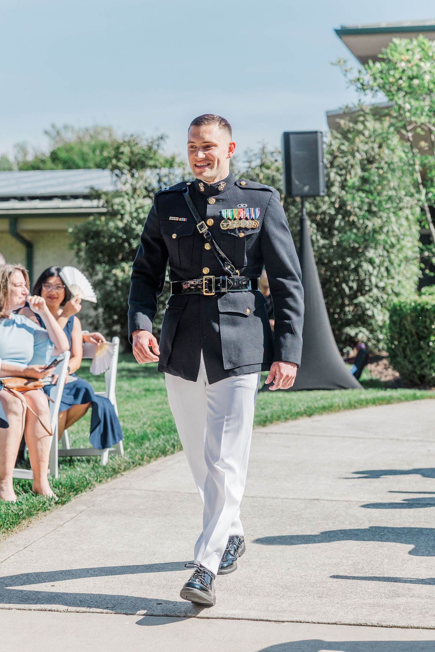 Smiling military officer in dress uniform with medals walks down outdoor aisle at a sunny wedding ceremony.