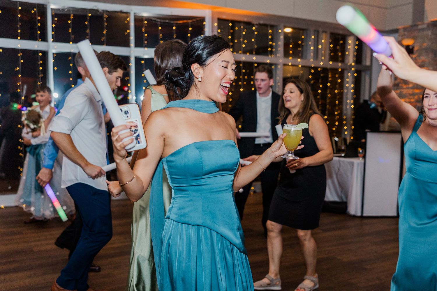 Woman in teal dress dancing joyfully at wedding reception with glow sticks and guests celebrating in background.