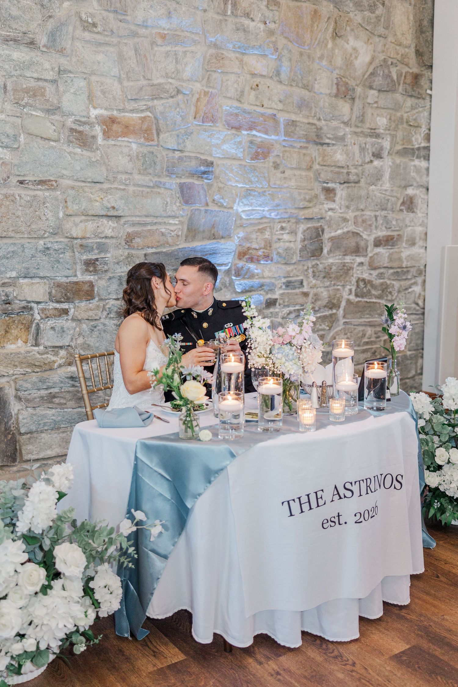 Newlyweds share a kiss at their sweetheart table decorated with candles and florals, featuring a custom The Astrivos est. tablecloth.