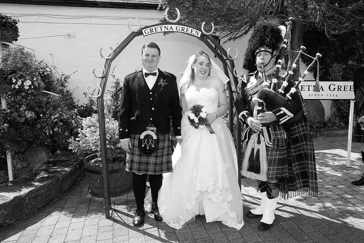 Bride and groom pose under floral arch at Gretna Green with bagpiper in traditional Scottish kilt attire.