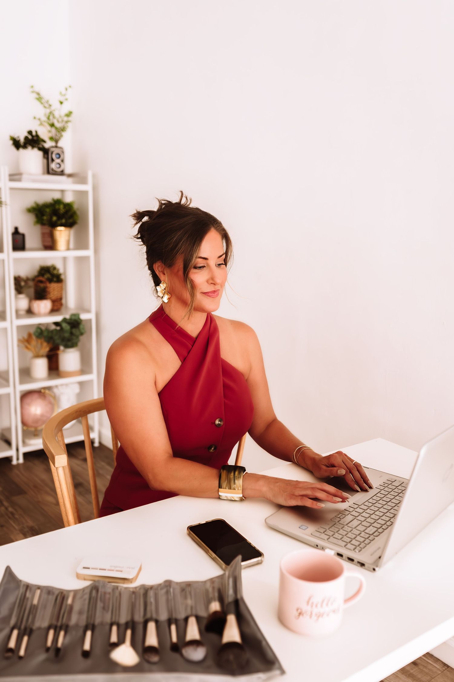 Brunette woman in red halter dress working on laptop at white desk with makeup brushes nearby.