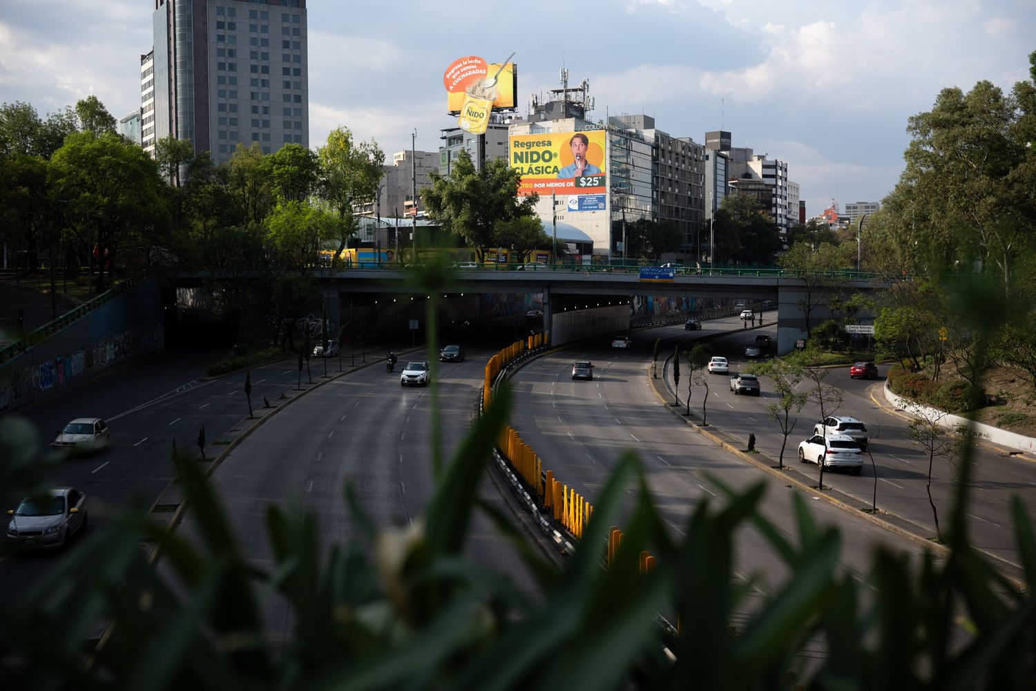 Aerial view of a busy multi-lane highway with cars, a green overpass bridge, and city billboards in Mexico City.