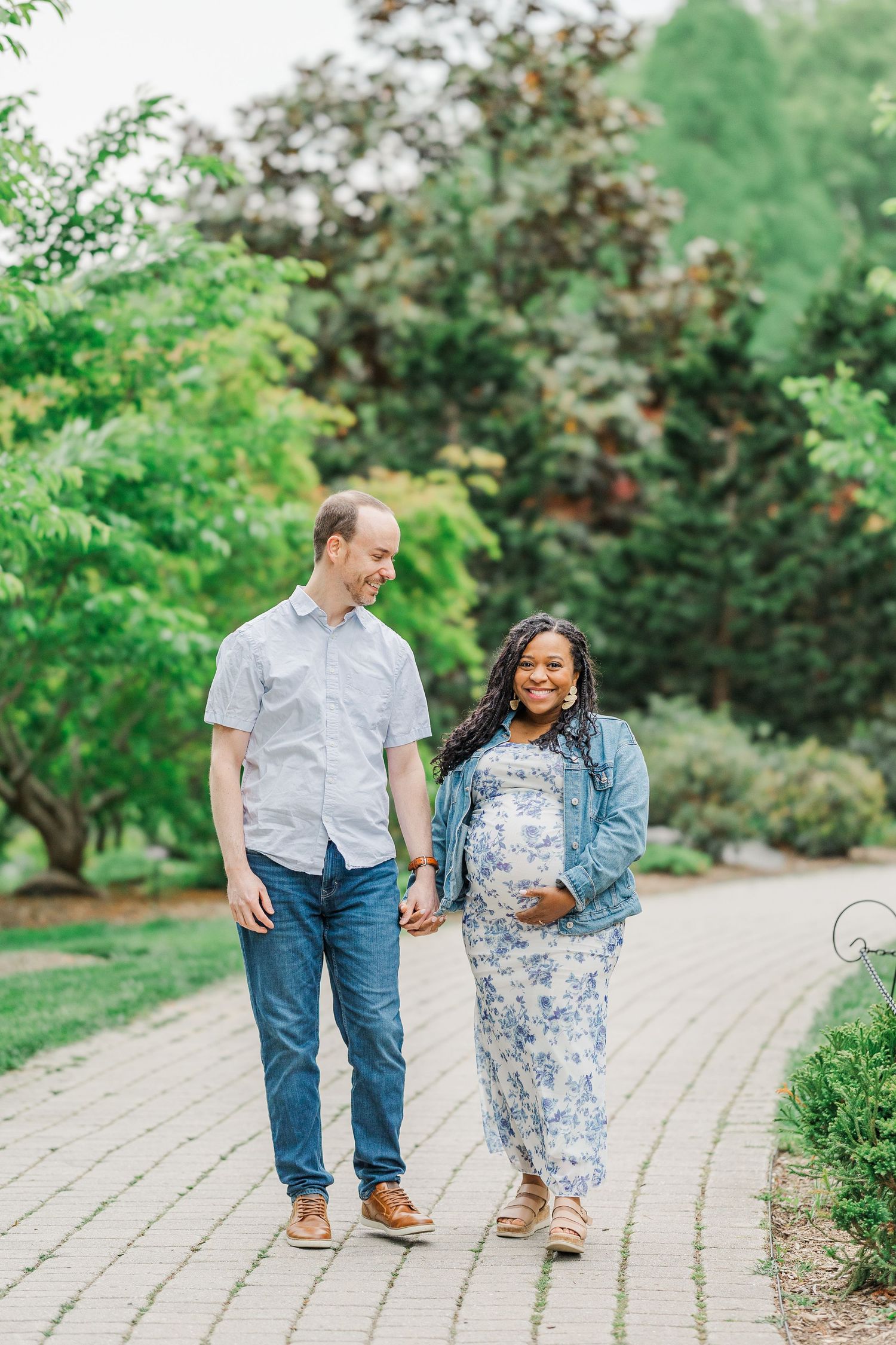 Pregnant couple holding hands walking on a brick path in a lush green garden, woman in floral dress smiling.