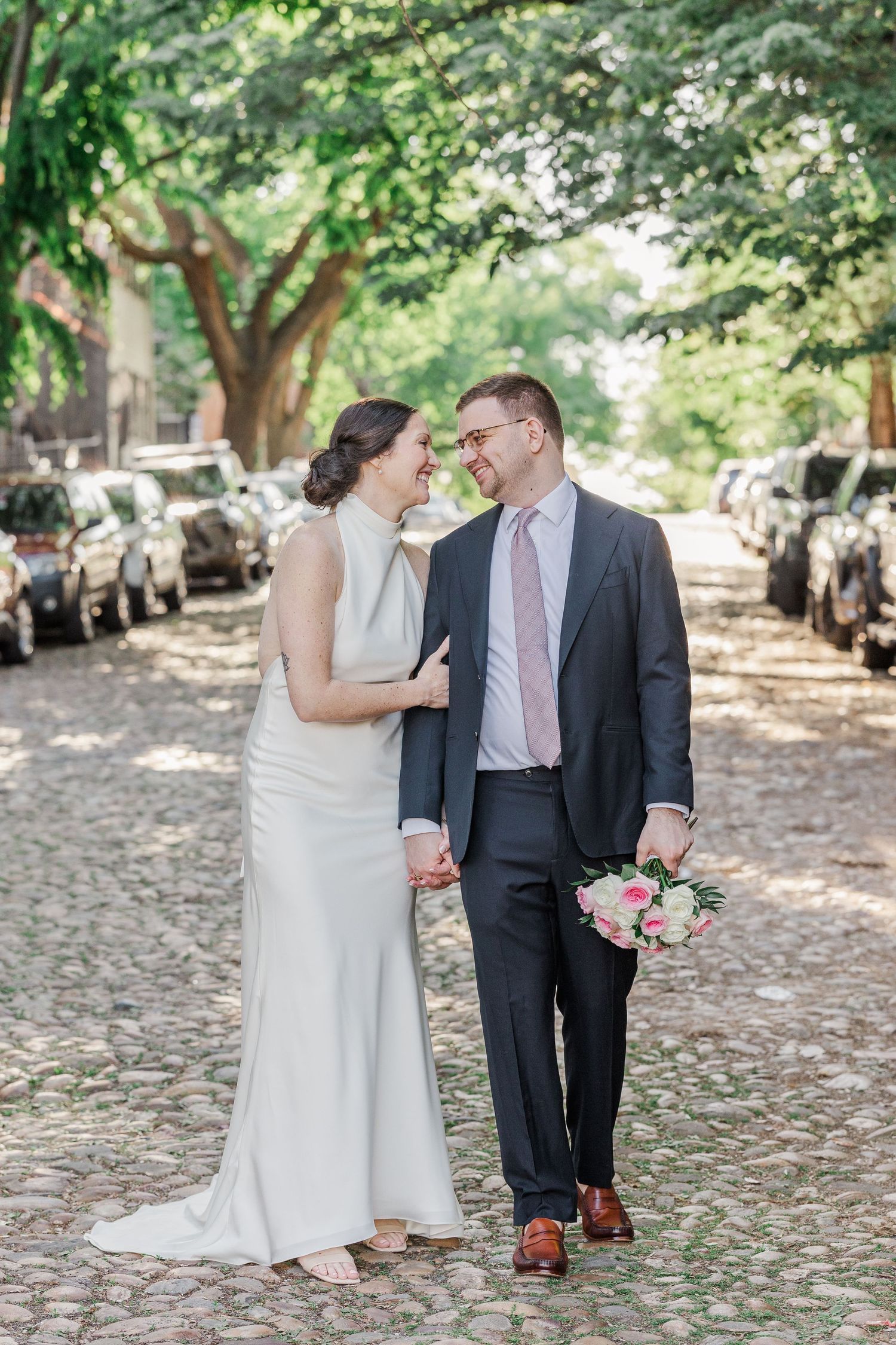 A bride and groom walk hand-in-hand on a cobblestone street, smiling at each other, with parked cars and trees in background.