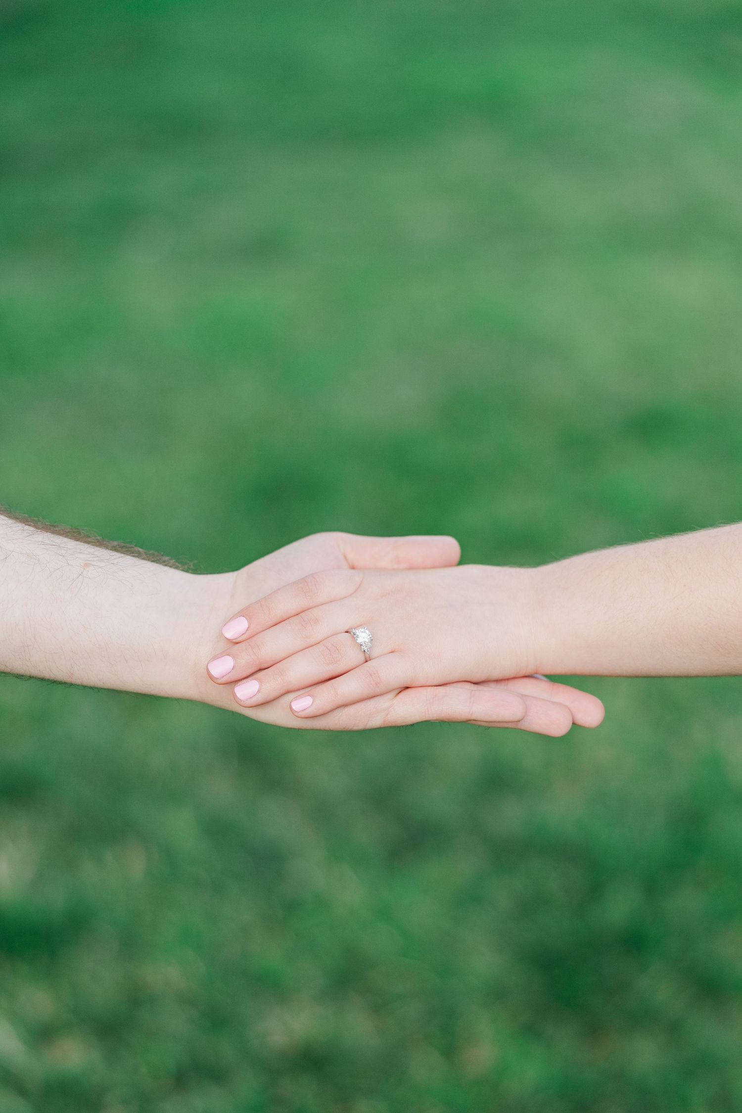 Two hands gently touching outdoors, one wearing a diamond engagement ring, with a blurred green grass background.