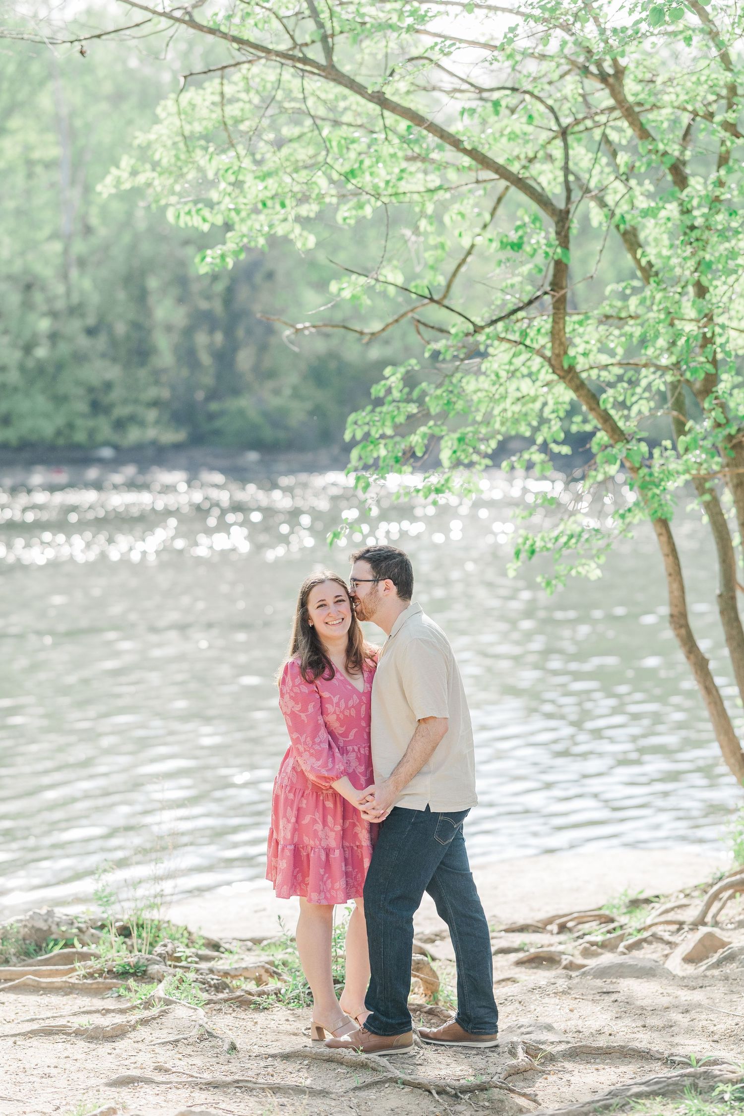 Couple shares a romantic kiss by a sunlit river, woman in pink dress smiling, surrounded by lush green trees.