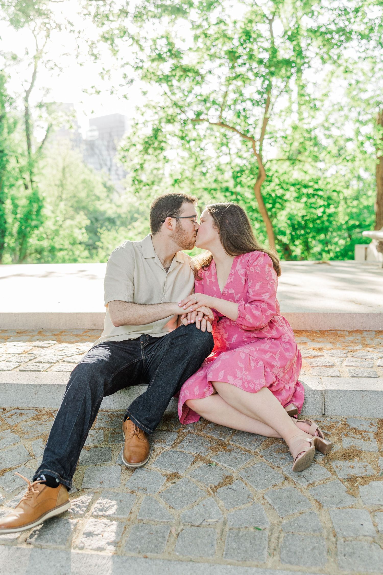 Couple sharing a kiss while sitting on stone steps surrounded by lush green trees in an outdoor park setting.