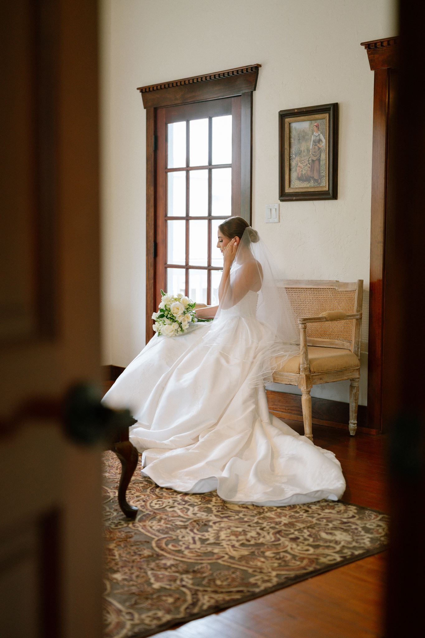 Bride in white gown with veil sits on bench holding bouquet near window in elegant room.